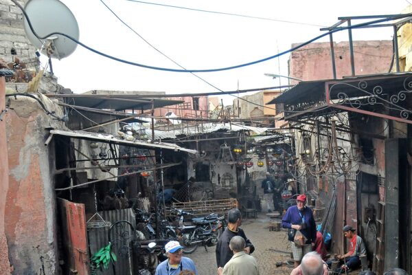 A narrow lane in the Marrakech medina lined with metalworkers' workshops, lanterns hanging under tin awnings and visitors standing among shopkeepers
