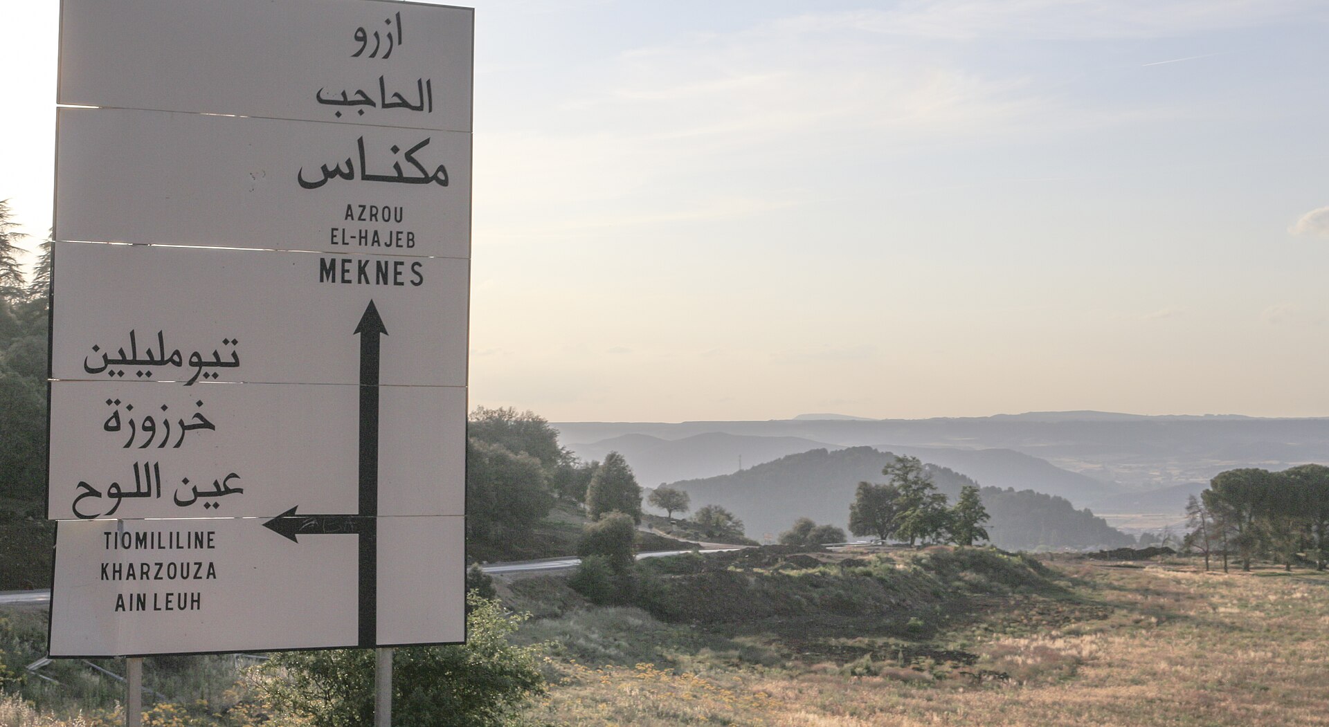 A white road sign near Azrou listing Azrou, El Hajeb, Meknes, Tiomililine, Kharzouza and Ain Leuh in Arabic script and French, with a forested hillside behind