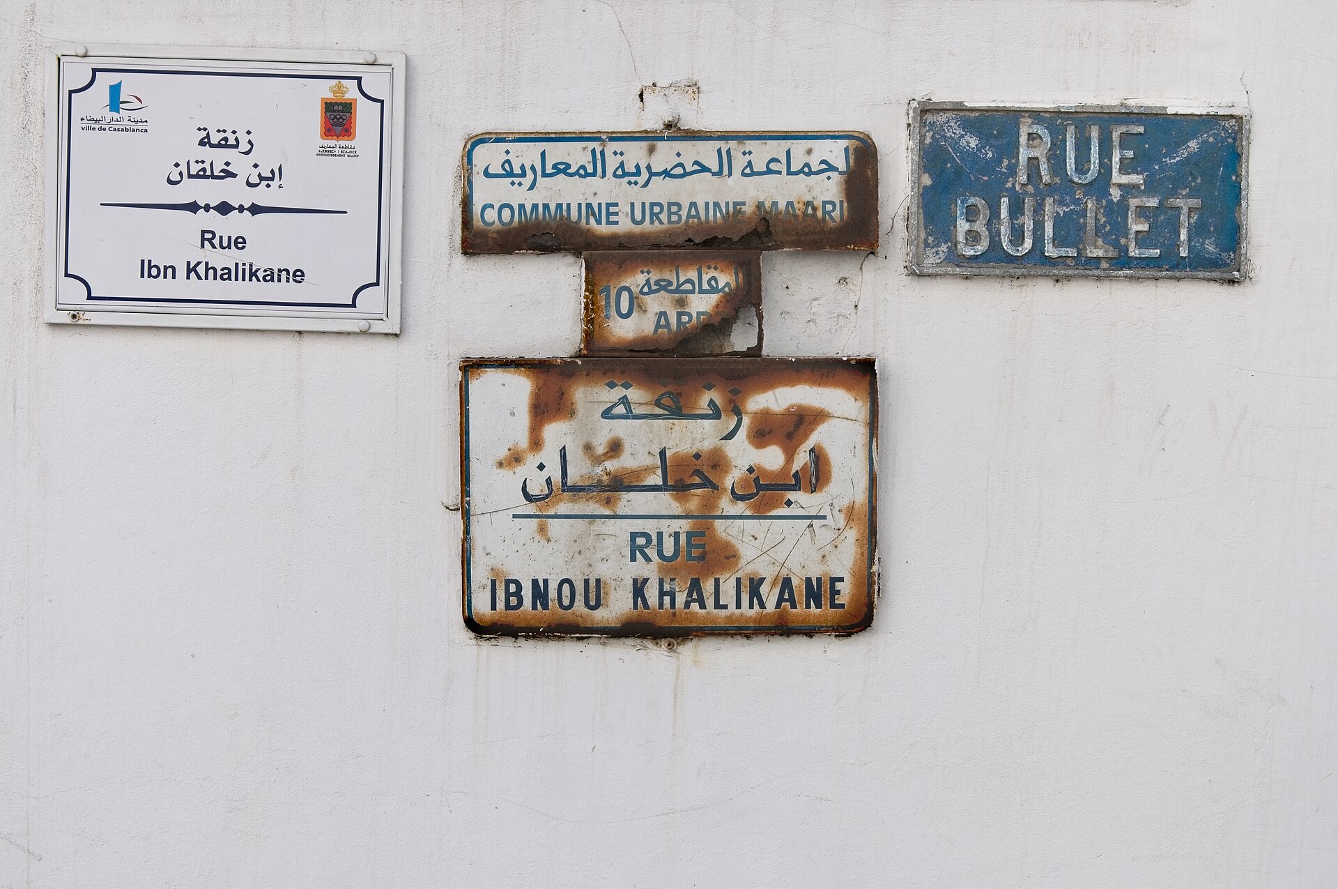 Old and new street name plates on a white wall in Casablanca: a blue French-only plate reading Rue Bullet, a rusted Arabic and French plate and a newer white Arabic and French plate reading Rue Ibn Khalikane