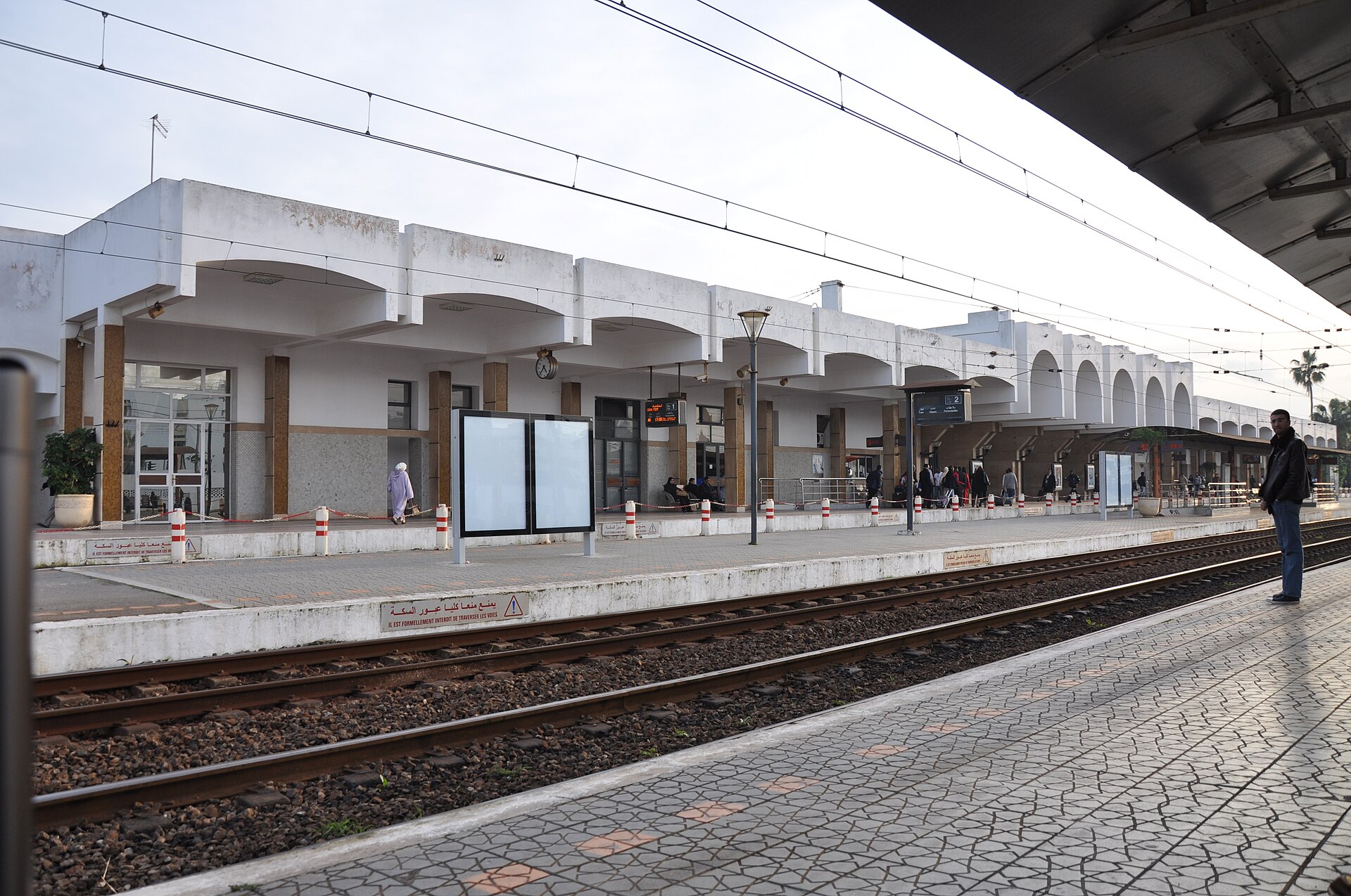 A low white railway station building with arches seen across the tracks from the platform, with a few passengers waiting and Arabic and French signs on the platform edge
