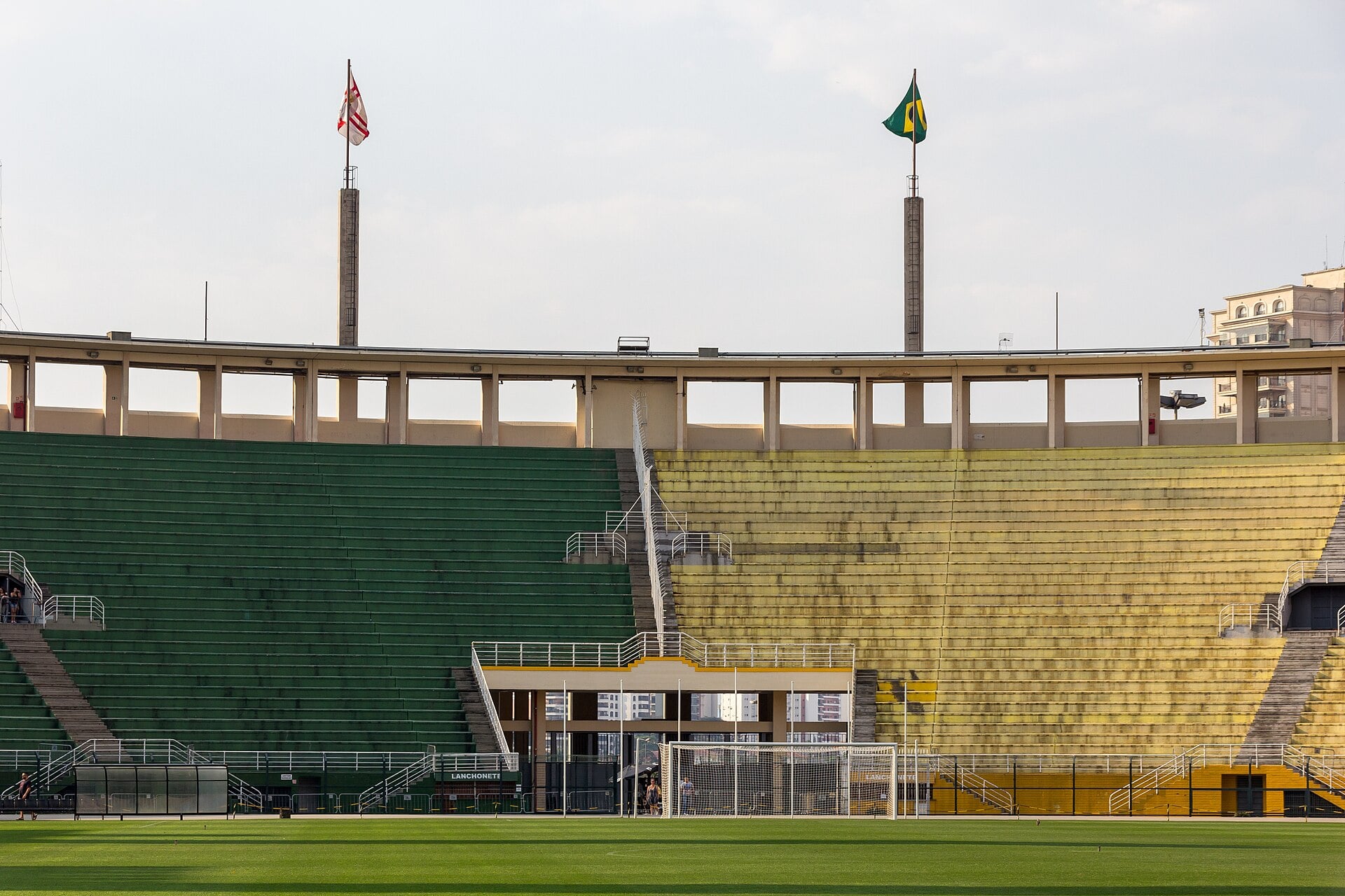 The green and yellow grandstands of the Pacaembu stadium in São Paulo, with a Brazilian flag and a São Paulo city flag on tall masts above the pitch.