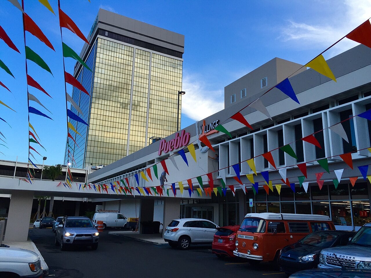 Entrance of a Pueblo supermarket at El Monte Mall in Hato Rey, San Juan, with colourful bunting, parked cars and an office tower behind