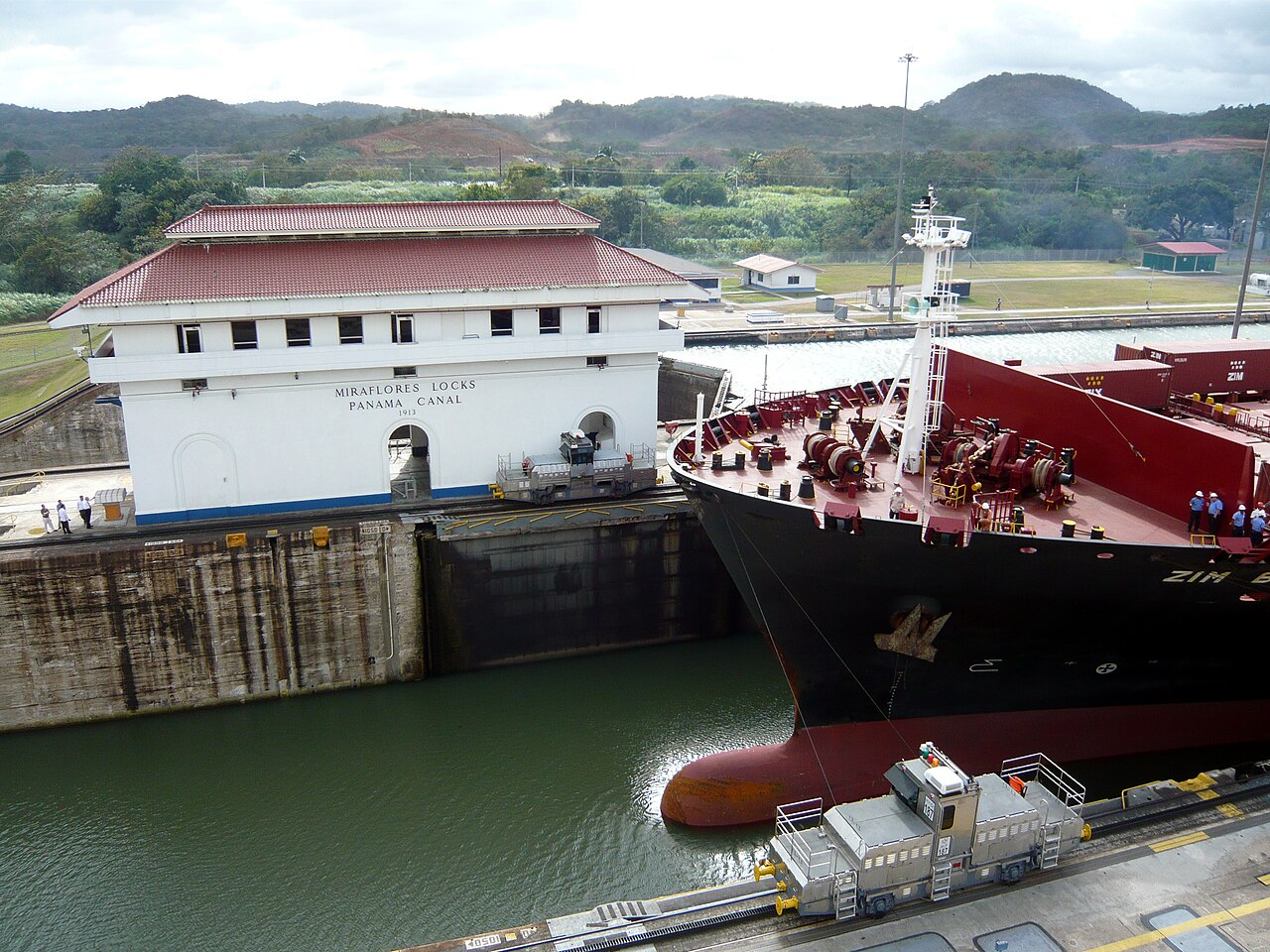 A container ship beside the Miraflores Locks building of the Panama Canal with a lock locomotive in the foreground
