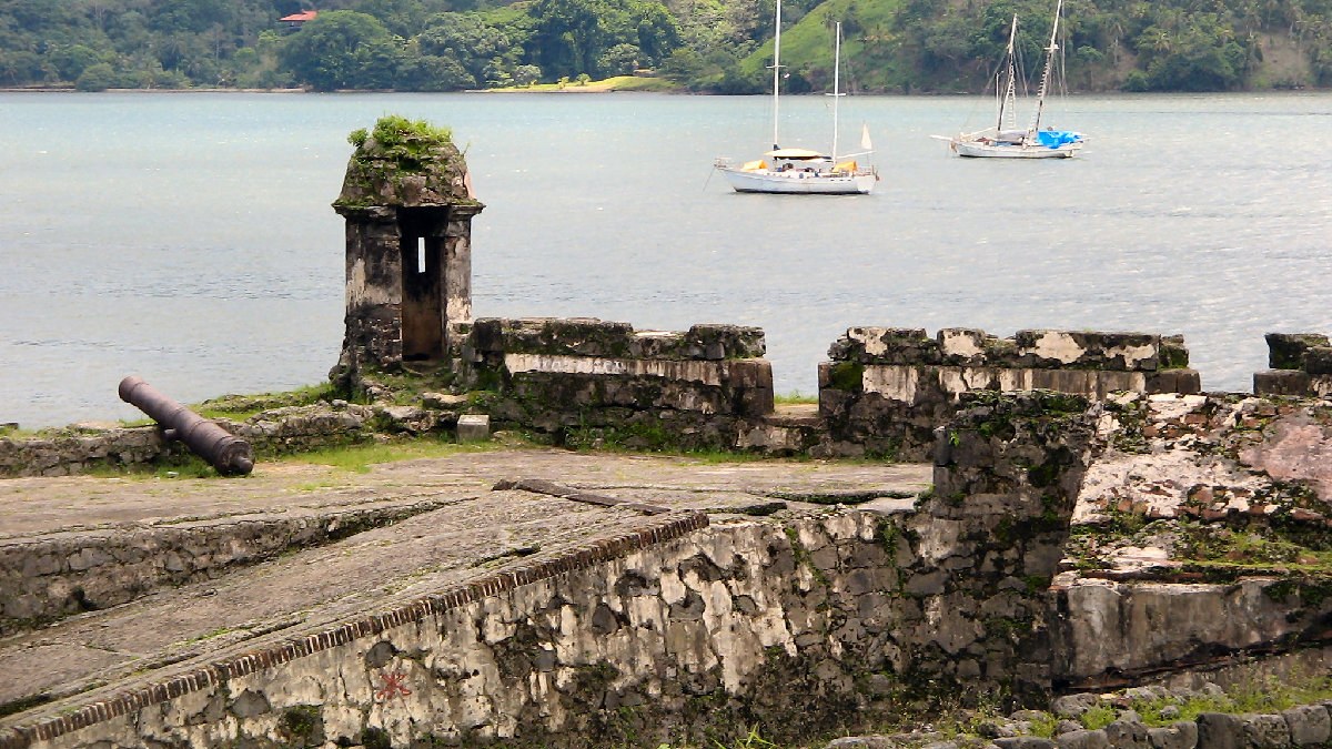 A stone sentry box, a cannon and fortress walls at Portobelo, Panama, with sailboats in the bay behind