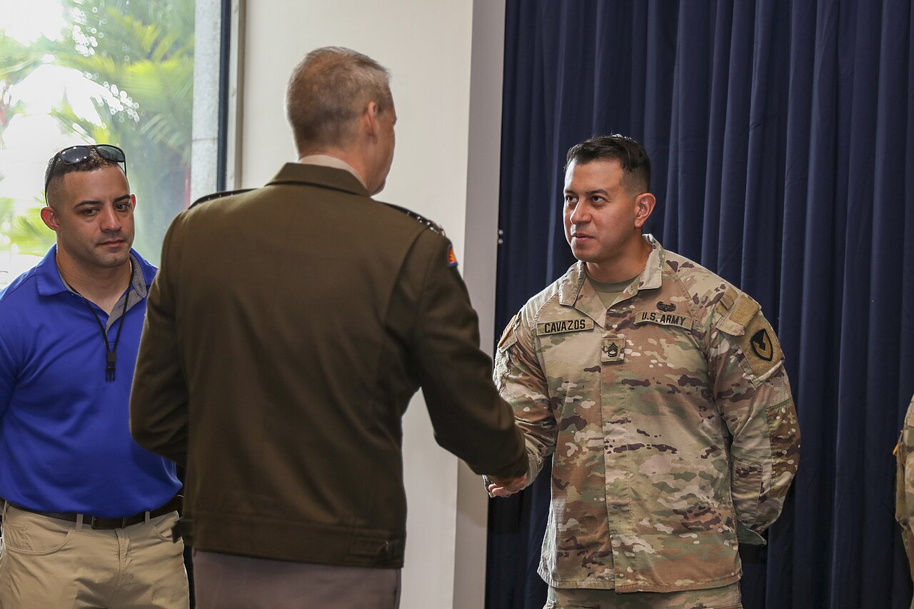 A US Army general, seen from behind in a brown uniform, shakes hands with a US Army soldier in camouflage beside a blue curtain