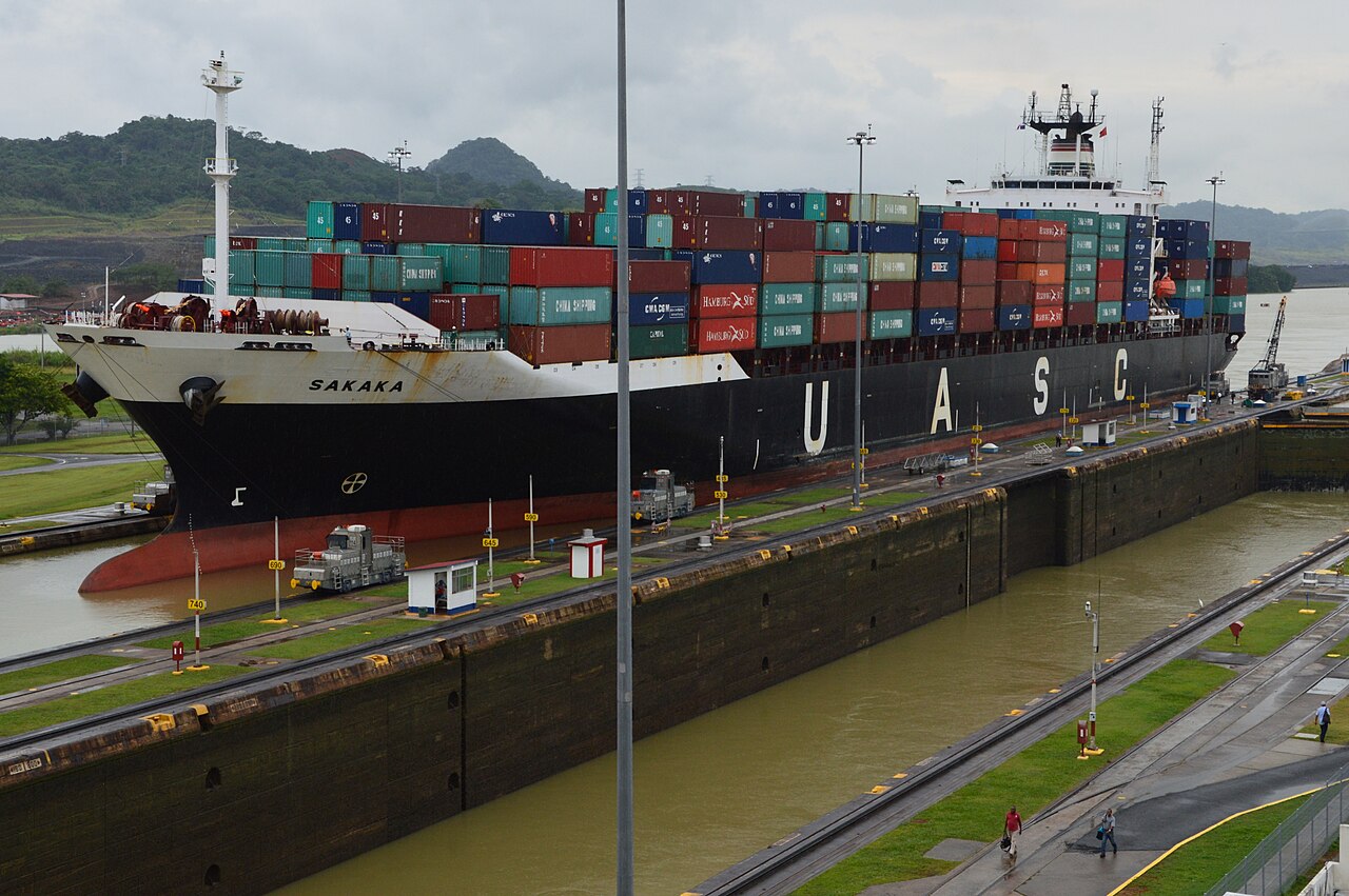 A large container ship loaded with colourful containers passes through a lock chamber of the Panama Canal, with locomotives on the lock wall and green hills behind.