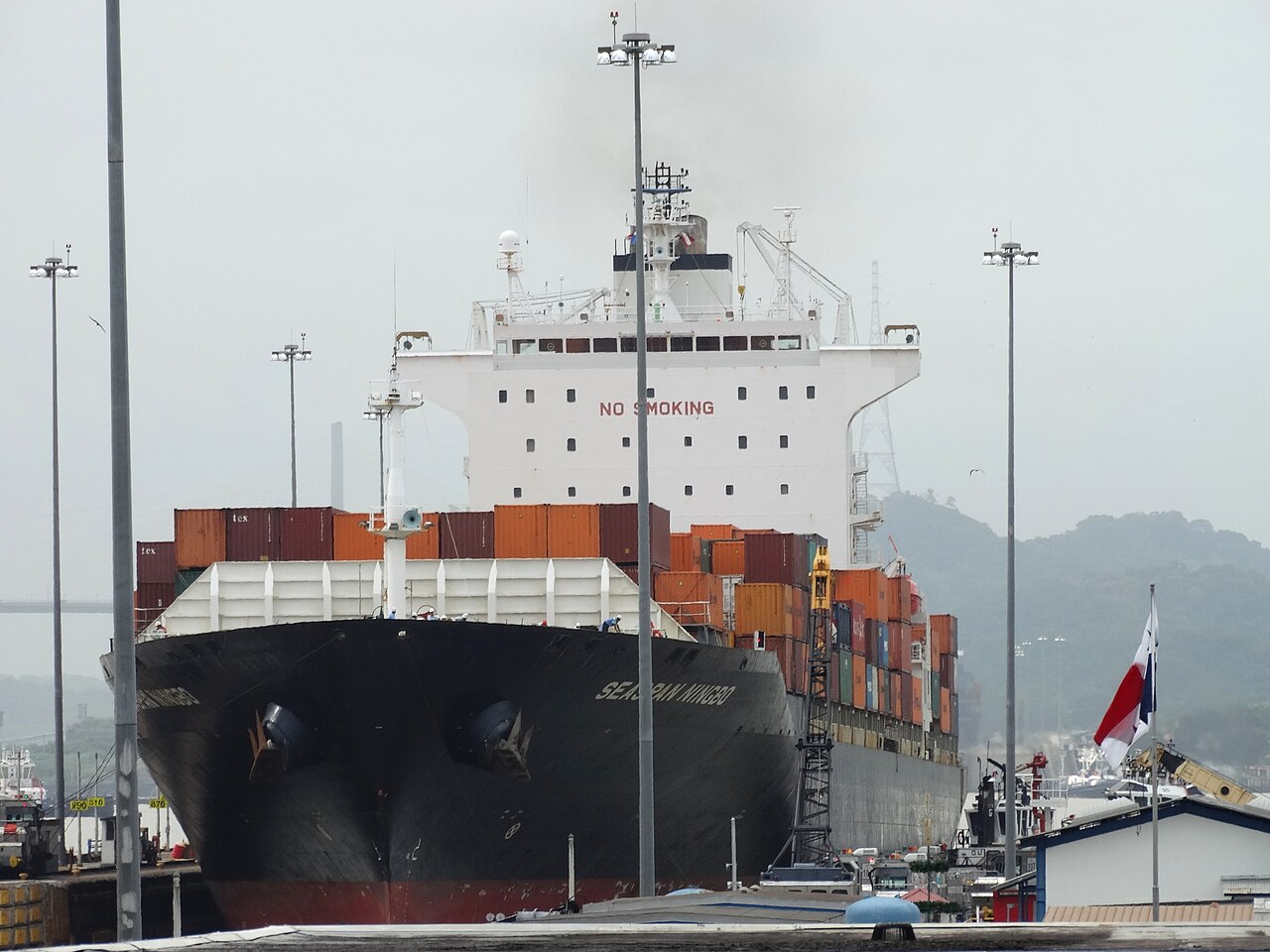 The bow of a black-hulled container ship stacked with containers, seen in a Panama Canal lock with a Panamanian flag at right.
