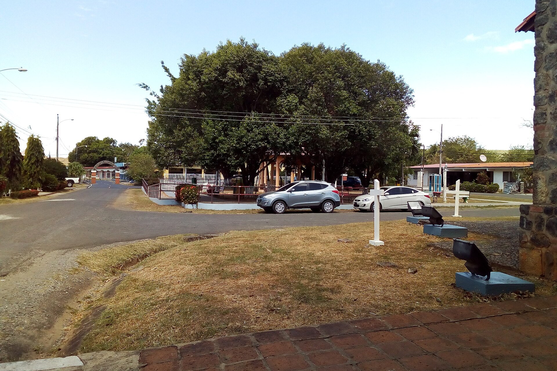 A small town park shaded by one large tree, with a stone wall and white crosses in the foreground, in San Francisco de la Montaña, Veraguas, Panama