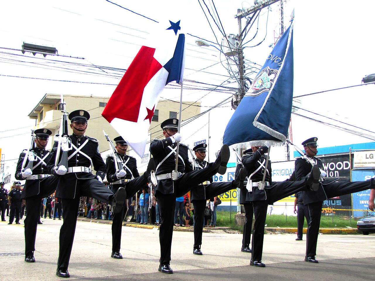 Panamanian police honour guard in dark dress uniforms marching with high kicks, carrying Panama's flag and a blue police banner