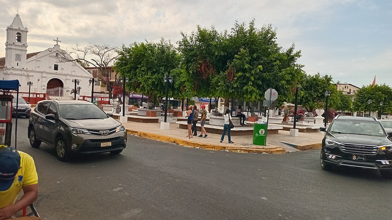 Belisario Porras Park in Las Tablas, Panama, with the white Santa Librada church on the left, trees, parked cars and pedestrians