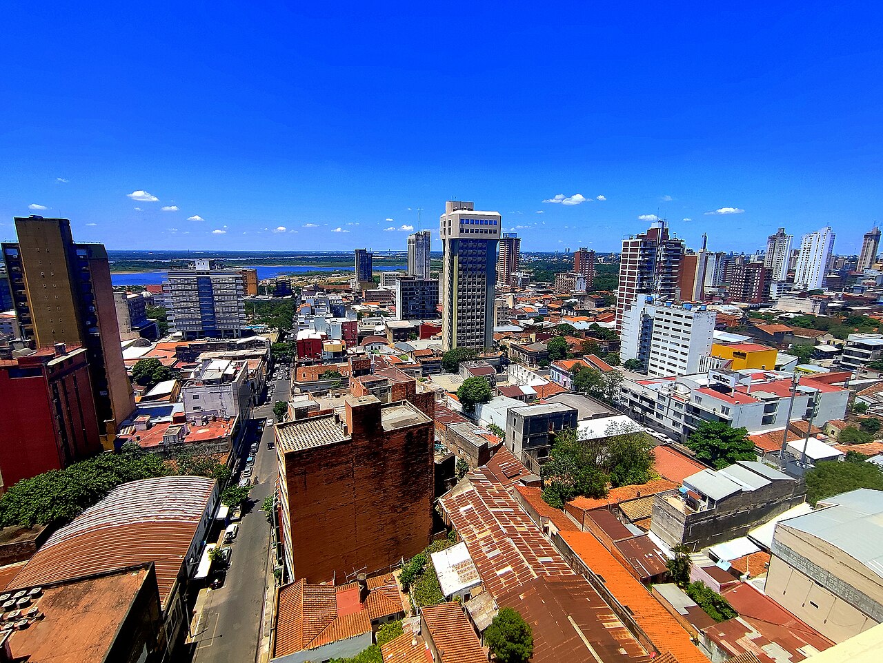 Rooftops and office towers of downtown Asunción under a deep blue sky, with a wide river and green far bank in the distance