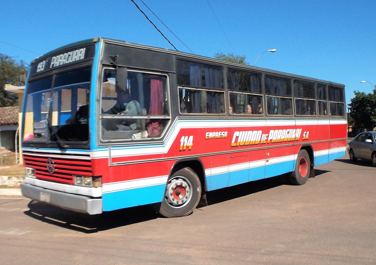 A red, white and blue bus of the Ciudad de Paraguarí company, route sign reading Paraguarí, driving on a paved road on a sunny day