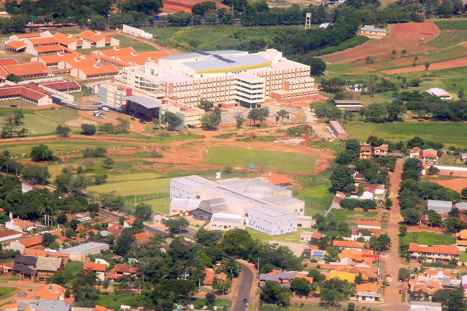 Aerial view of a large hospital complex with pale buildings and orange roofs among green fields and houses in San Lorenzo, near Asunción, Paraguay