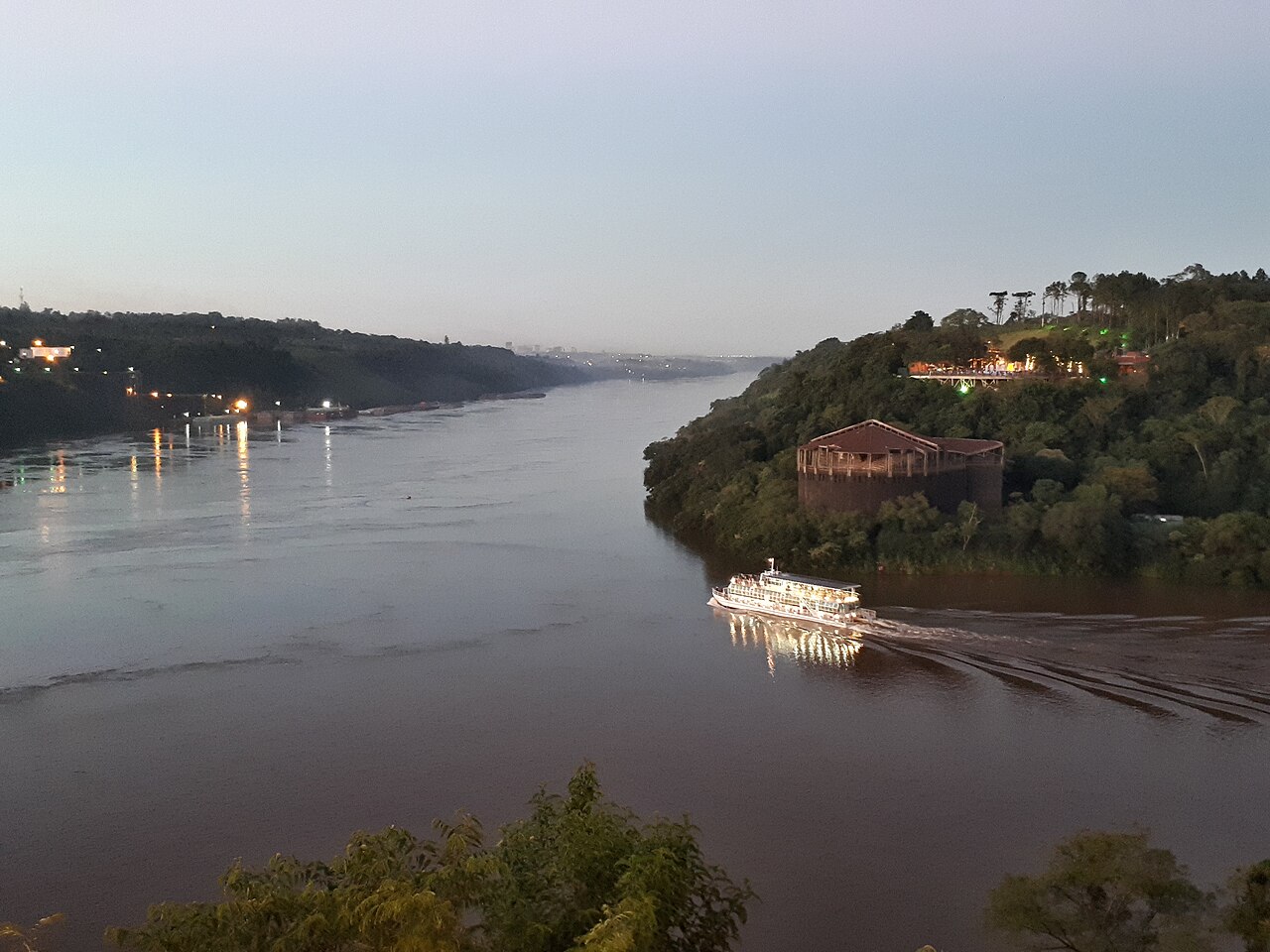 The wide brown Paraná River at dusk where the Iguazú River joins it, with a lit tour boat and forested banks on both sides