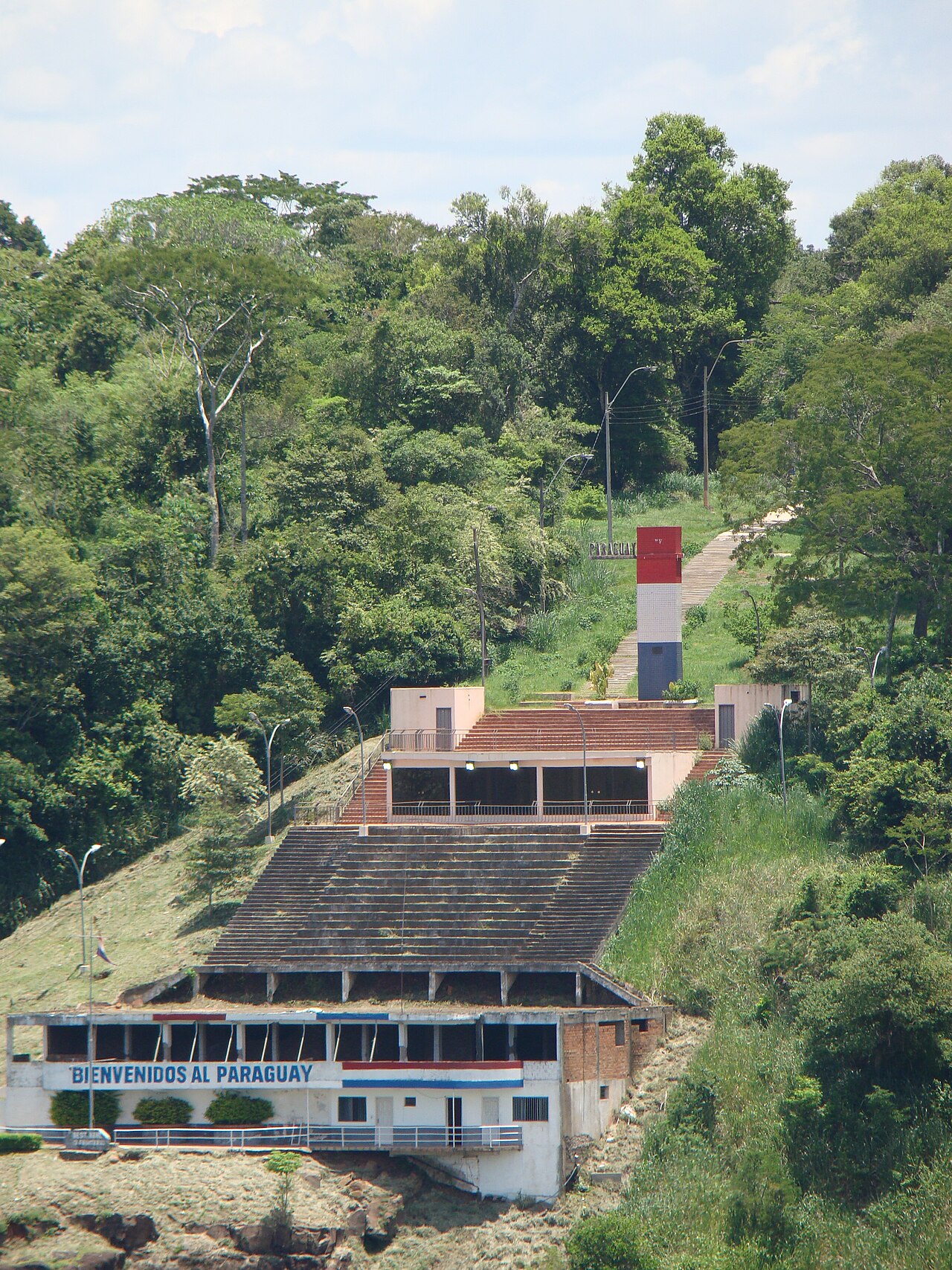 A white riverside building reading Bienvenidos al Paraguay below a red, white and blue obelisk on a forested hillside