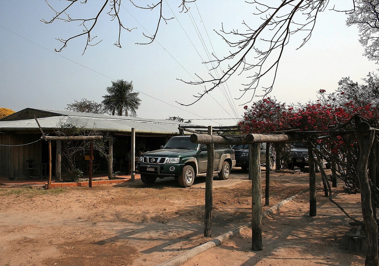 Single-storey ranch buildings with a corrugated metal roof, pickup trucks under a wooden pergola and red bougainvillea at an estancia in Paraguay's Chaco
