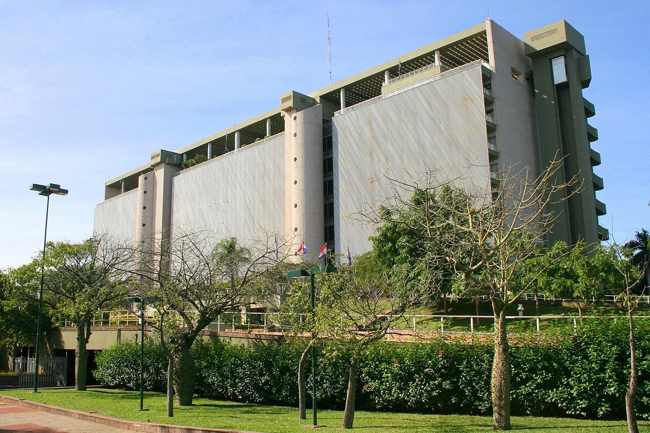 The Banco Central del Paraguay headquarters, a tall grey and cream concrete building with long vertical panels, seen across a lawn with trees and national flags