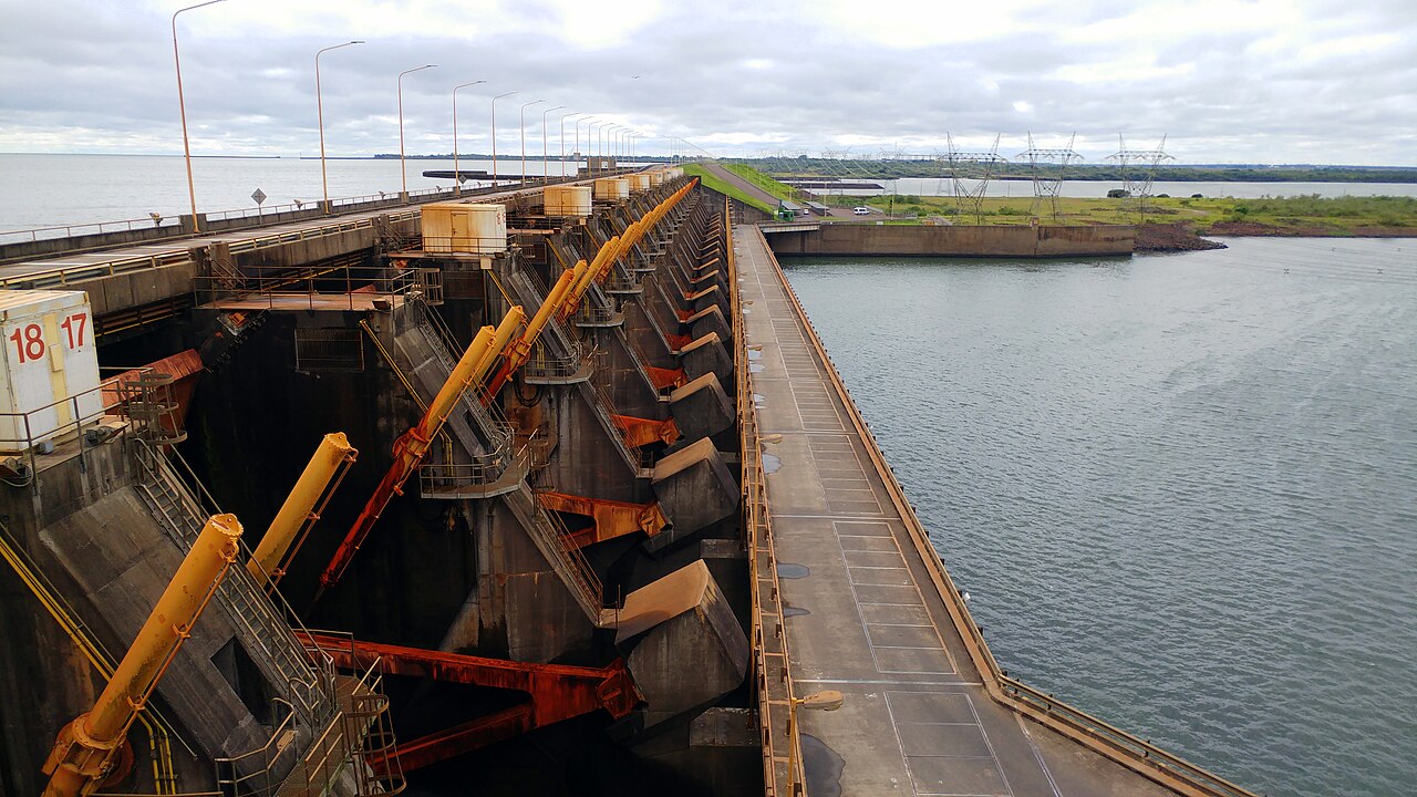 Spillway of the Yacyretá dam with yellow gate arms beside a wide reservoir and power pylons on the far bank