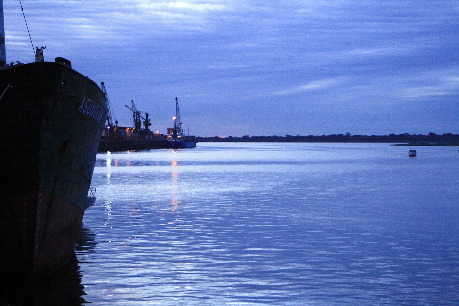 A cargo ship docked on the Paraguay River at dusk, with port cranes in the background