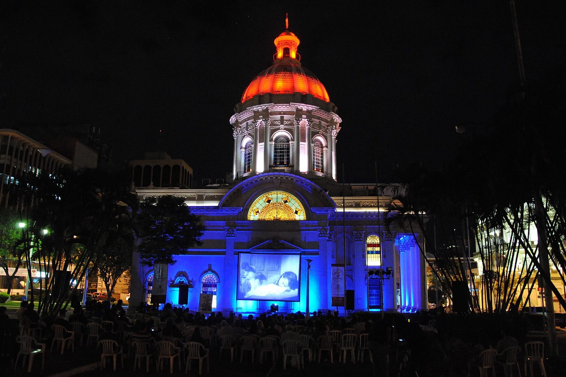 The domed National Pantheon of the Heroes in Asunción lit in blue and orange at night, with an audience seated on the lawn watching an outdoor film screening