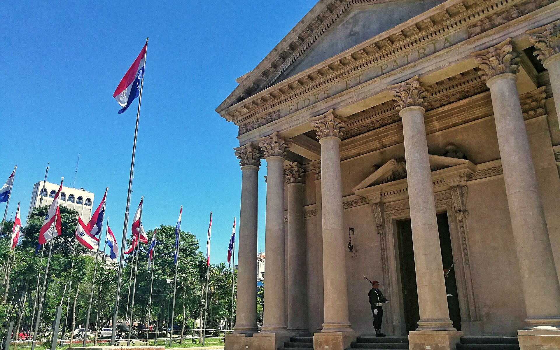 An honour guard in a red beret stands at the columned entrance of the National Pantheon of the Heroes in Asunci&oacute;n, with Paraguayan flags on the left under a blue sky