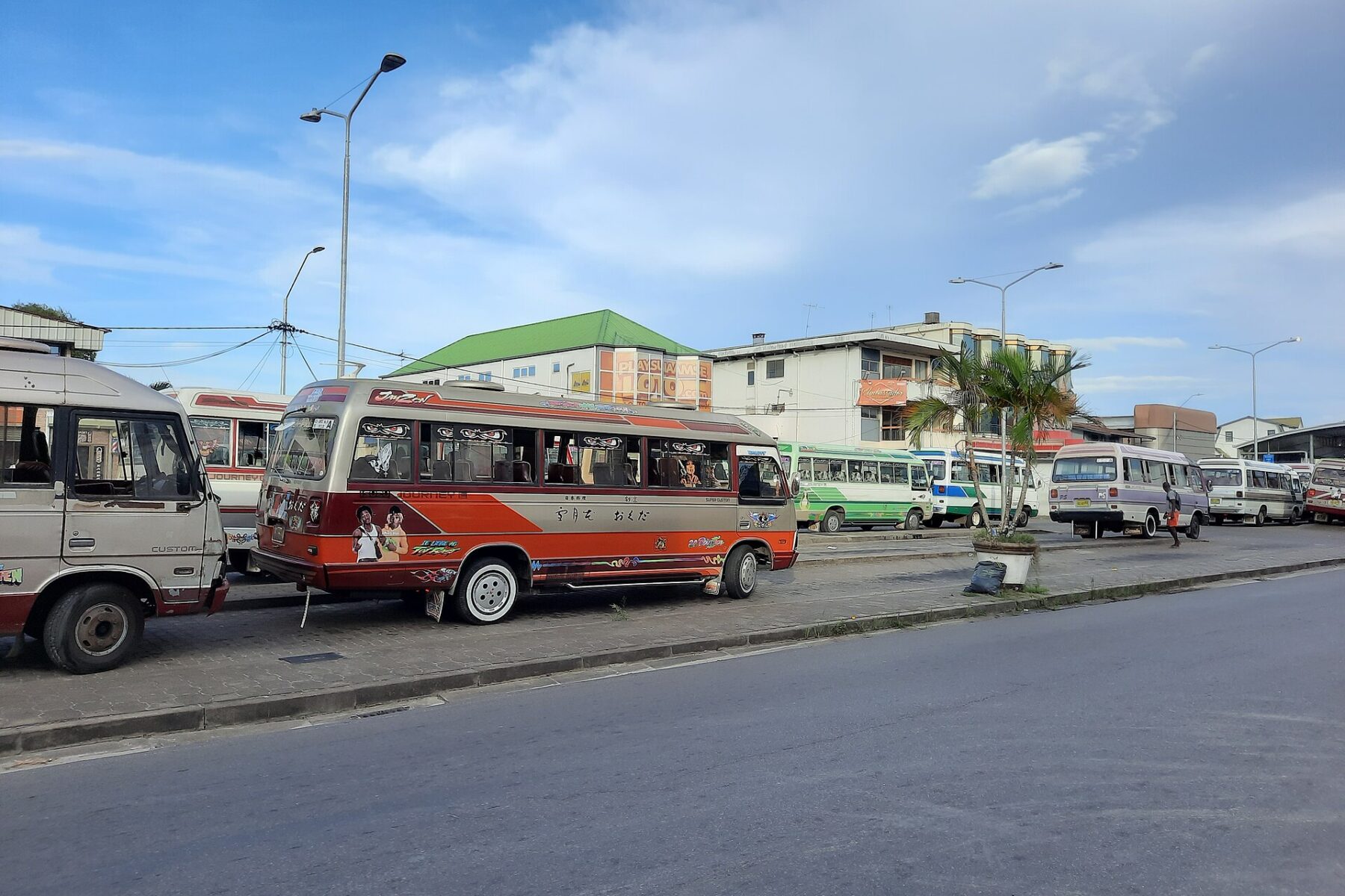 Privately run minibuses in bright paint parked in a row at a bus stand in Paramaribo, with a green-roofed building behind them