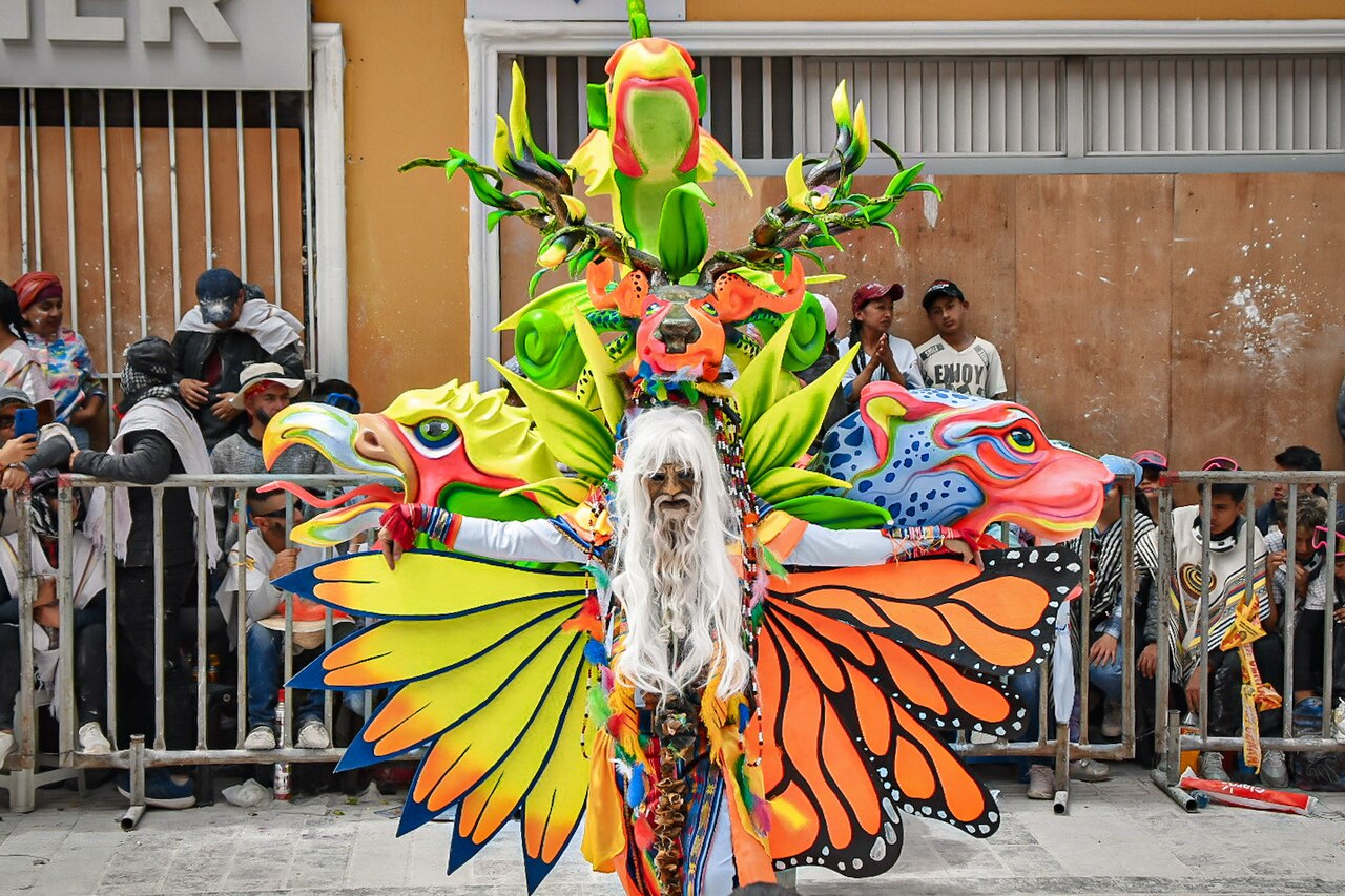 A costumed performer with butterfly wings and a white wig in the Desfile Magno parade of the 2024 Blacks and Whites Carnival in Pasto, Colombia, with spectators behind barriers.