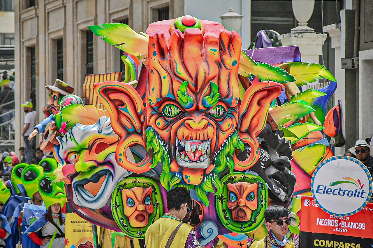 A large colourful carnival float with a snarling orange mask face rolling past a stone building in the Desfile Magno parade in Pasto, Colombia.