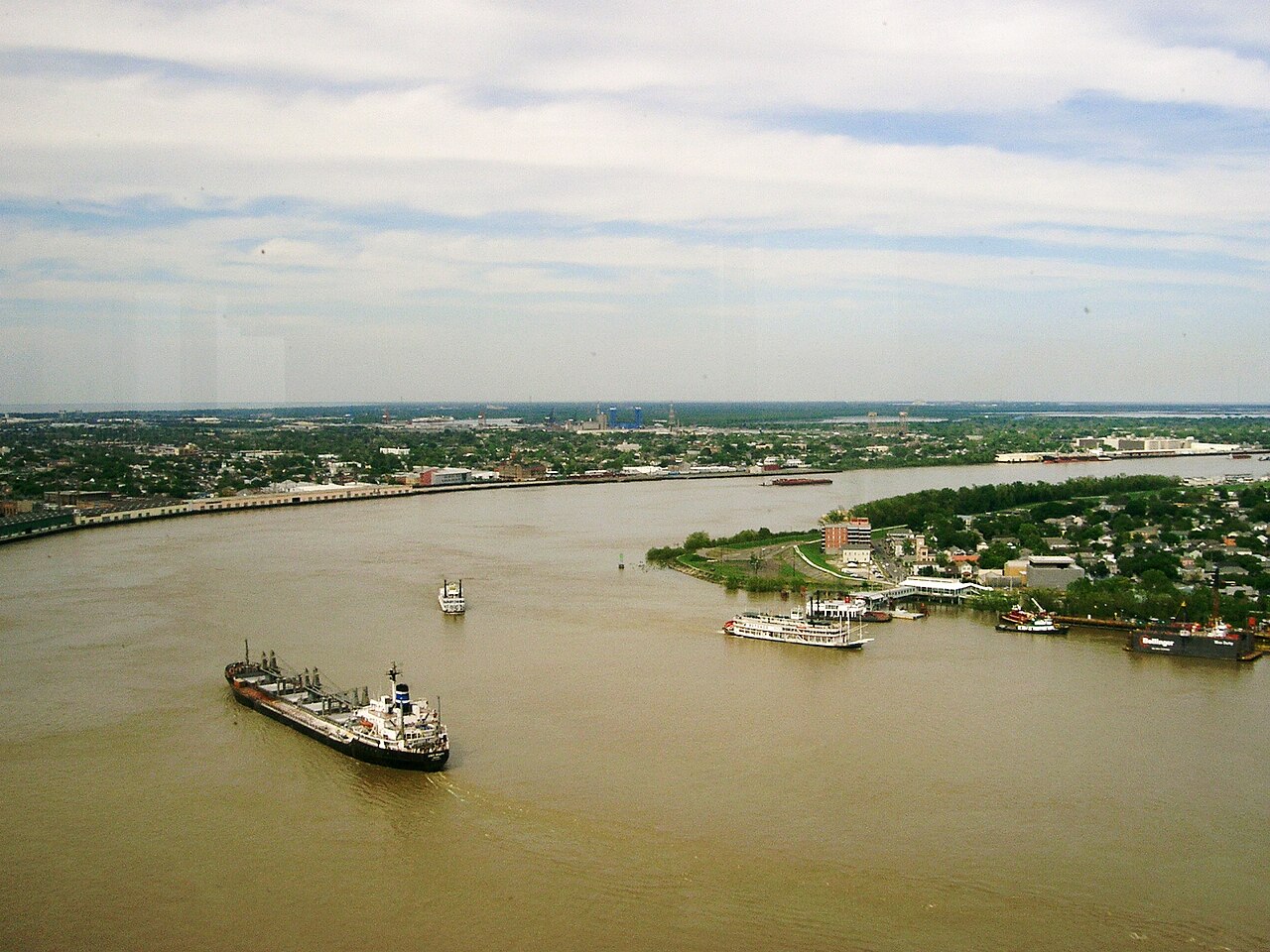 A cargo ship sails on the brown Mississippi River at New Orleans, with a paddle steamer and the Algiers Point shore behind