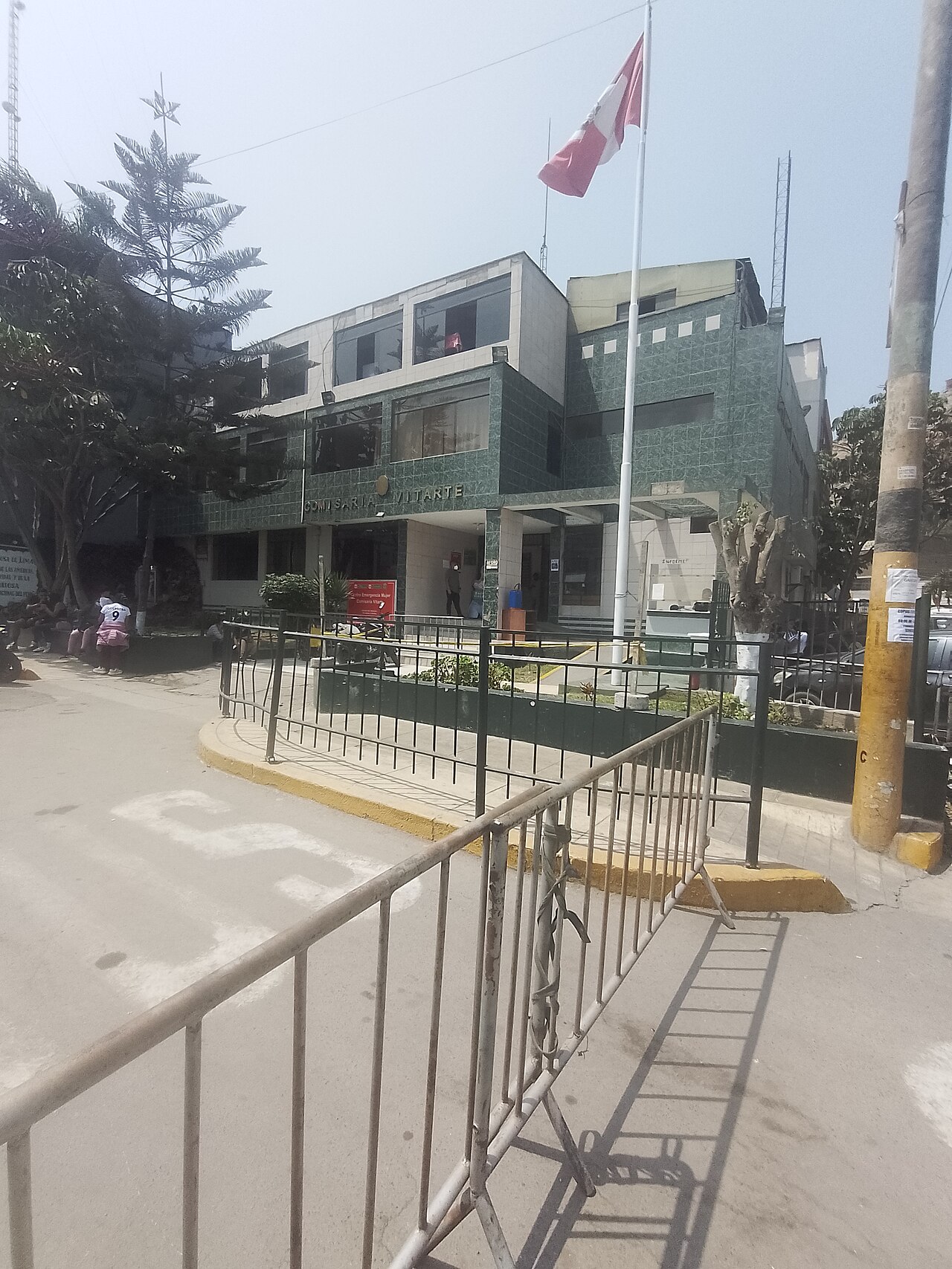 Three-storey police station with a green tiled facade and a Peruvian flag on a tall pole, behind street barriers, in Ate district, Lima