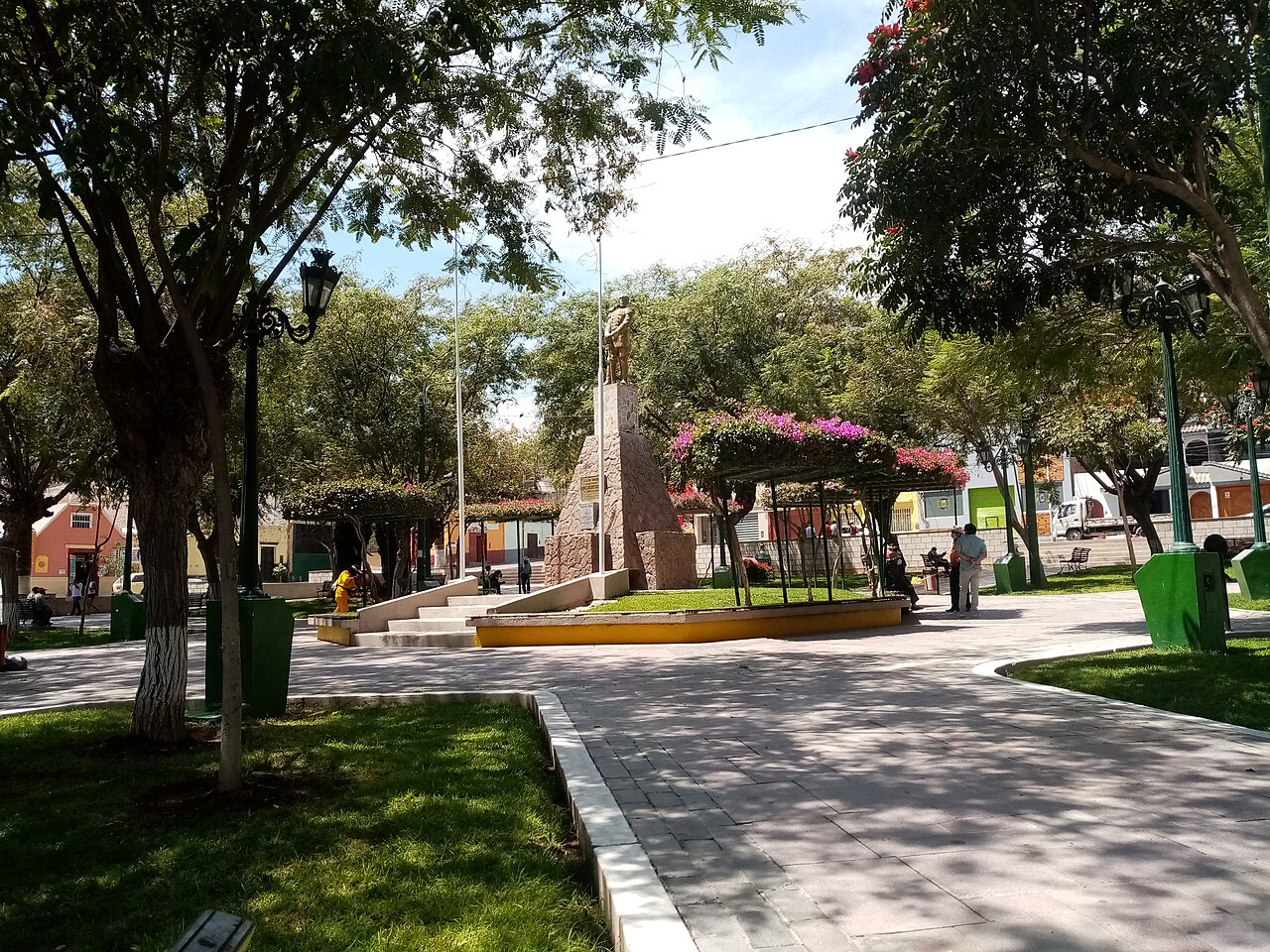 A shaded public square in the city of Moquegua with a monument, street lamps and flowering trees