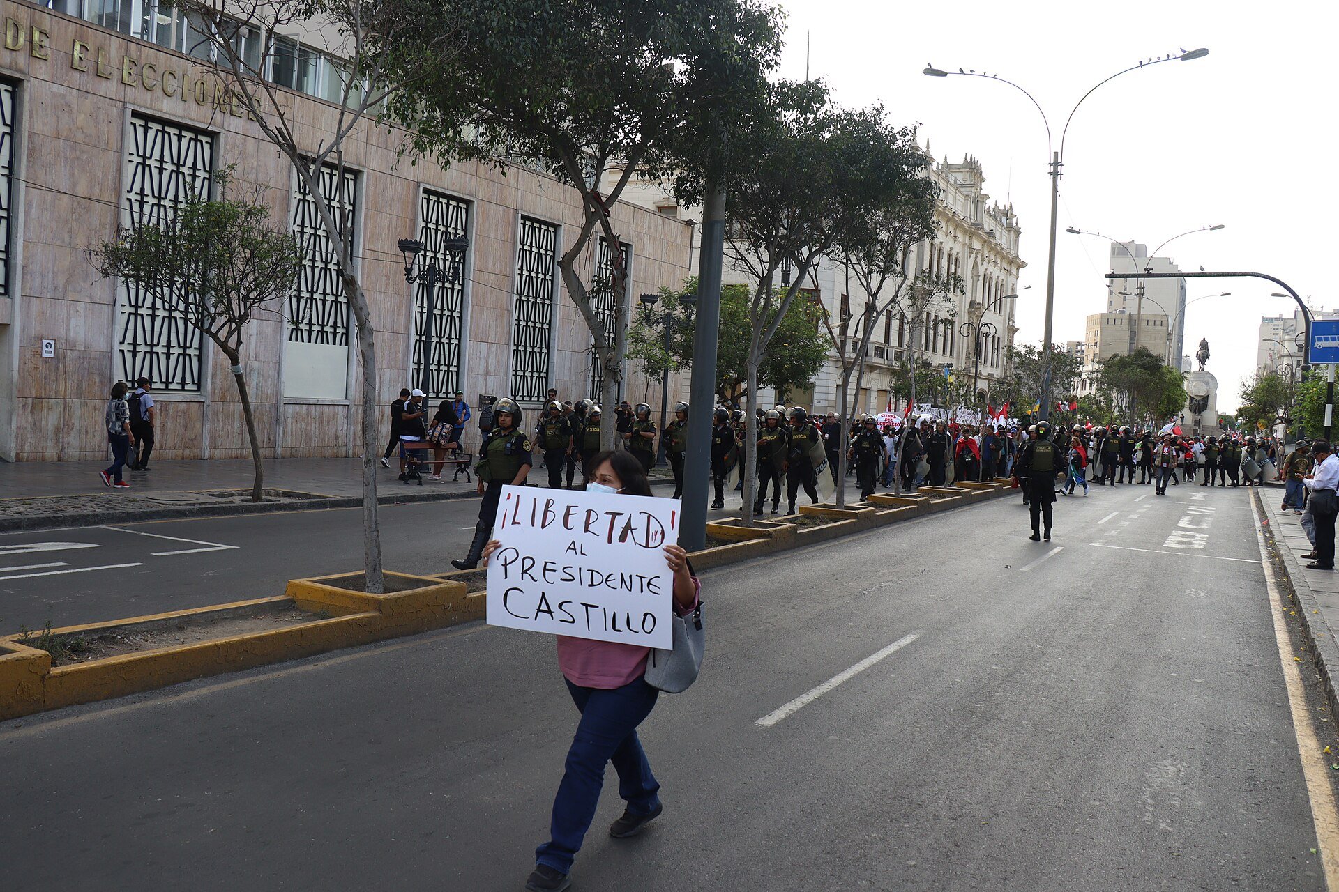 A woman walks down a Lima street holding a sign reading Libertad al Presidente Castillo, with police in riot gear and protesters behind her