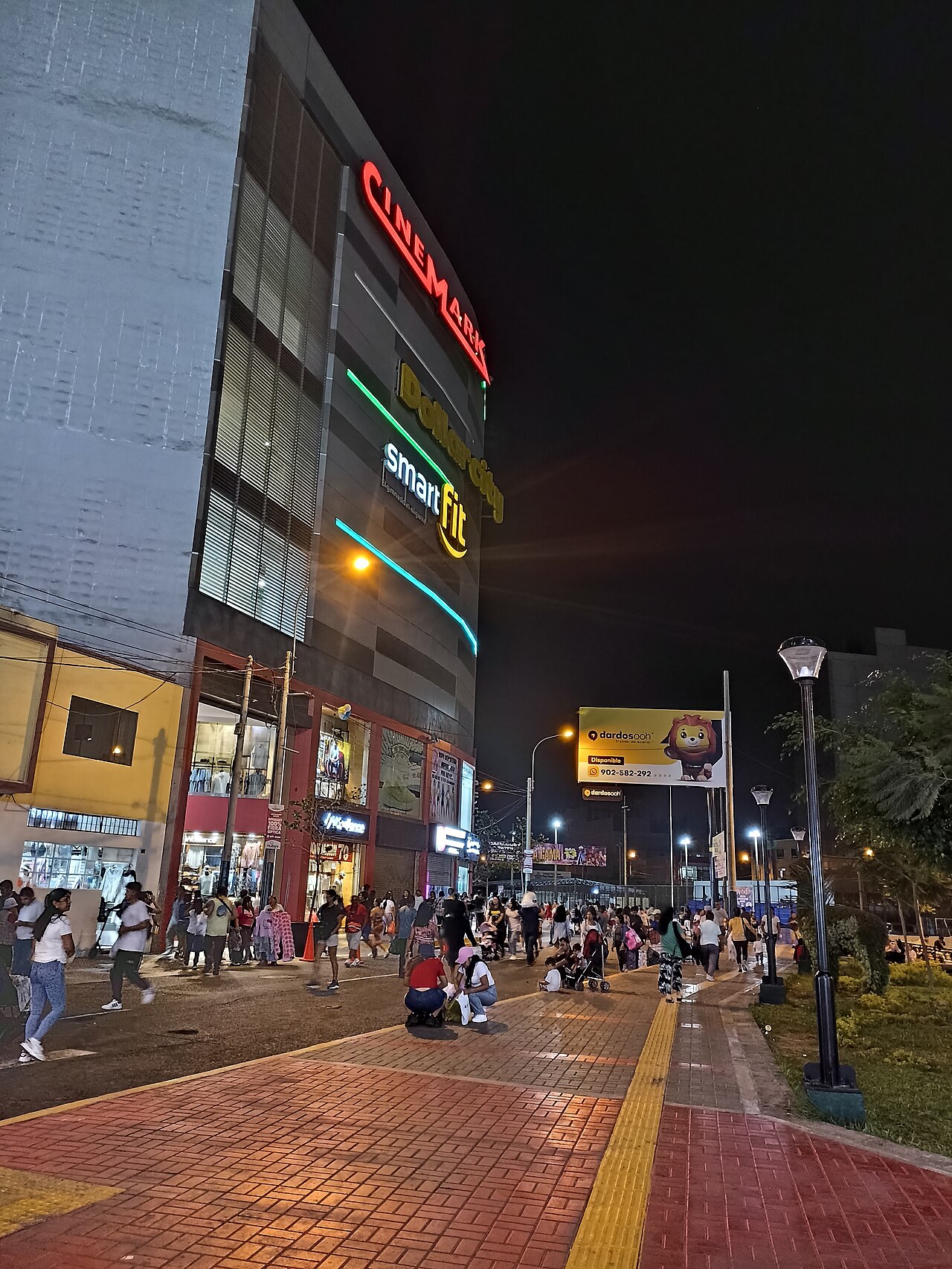 Shoppers walk past a lit commercial building with clothing shops at street level in Gamarra, Lima, at night