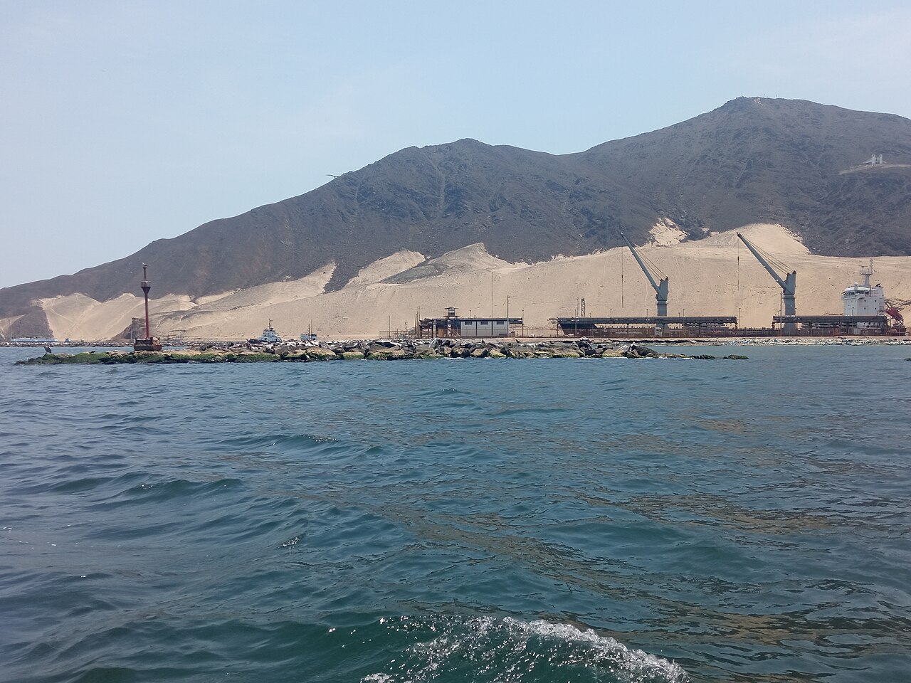 View from the sea of a port with two cranes, a docked cargo ship and a rock breakwater below sandy desert hills