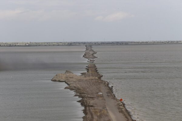 A road embankment running across a wide flooded plain in northern Peru, with brown floodwater on both sides and a warning sign on the shoulder