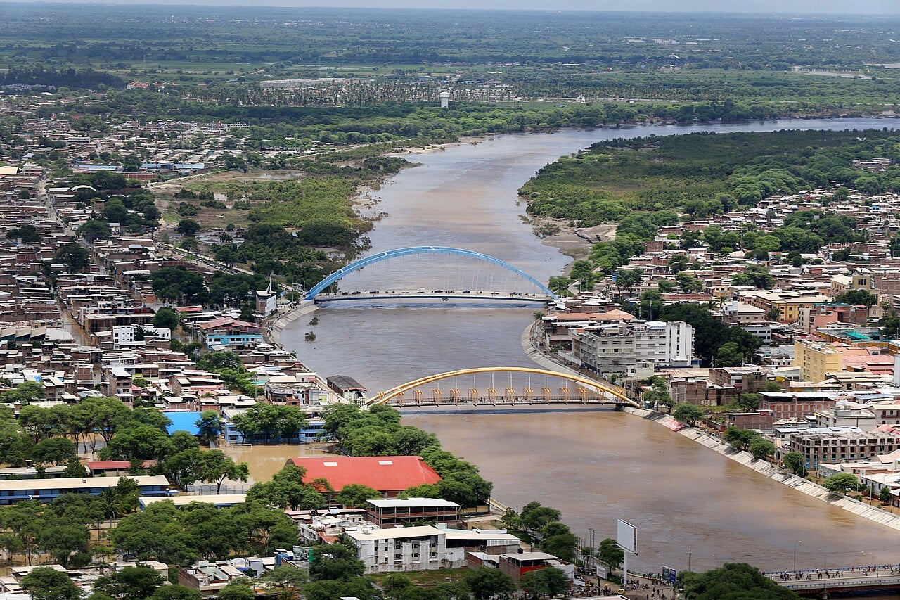 Aerial view of the Piura River in flood, brown water running between two arched bridges and city blocks on both banks in northern Peru