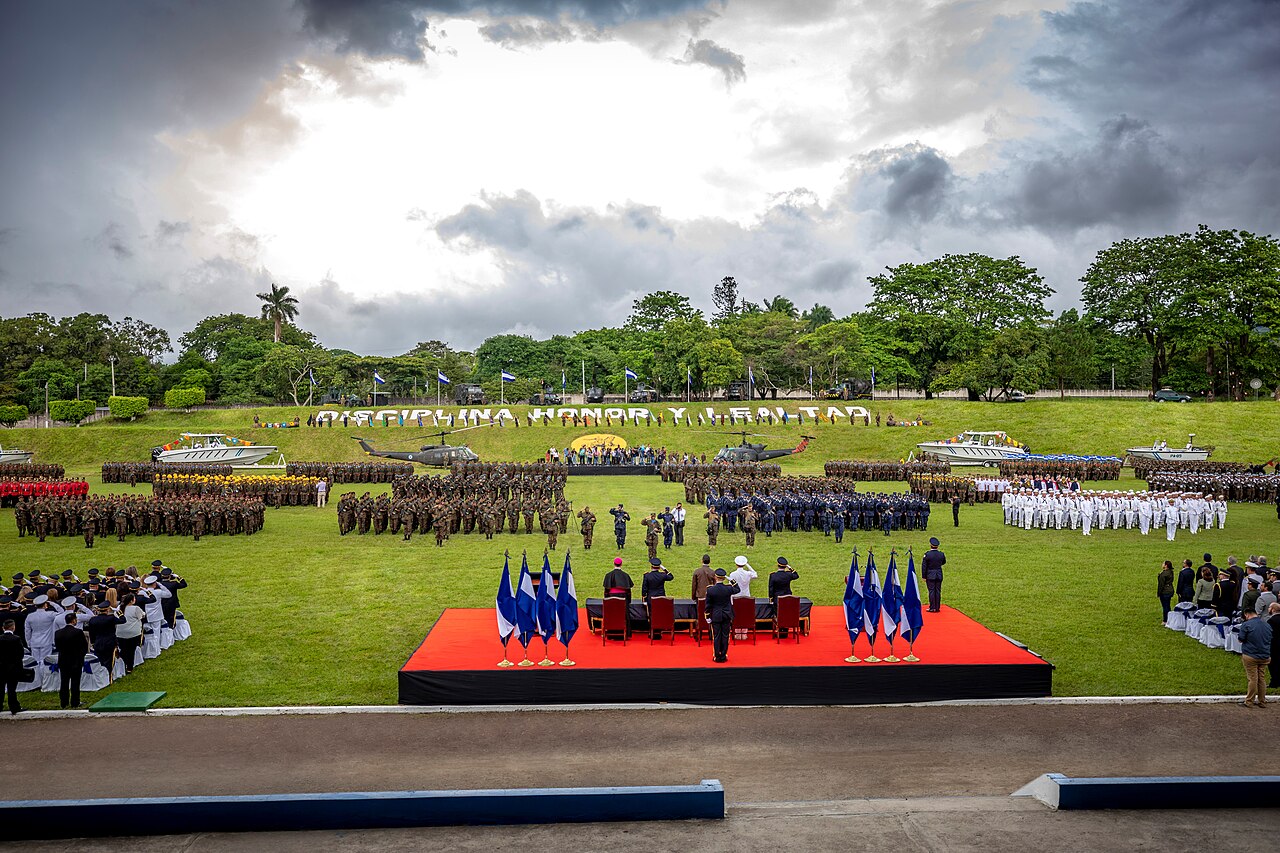 Soldiers, sailors and officers in formation on a green field at an armed forces ceremony in El Salvador, with helicopters and boats behind