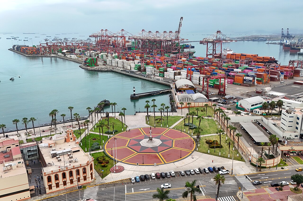 Aerial view of the port of Callao with red gantry cranes, stacked containers and ships at anchor beyond a seafront plaza