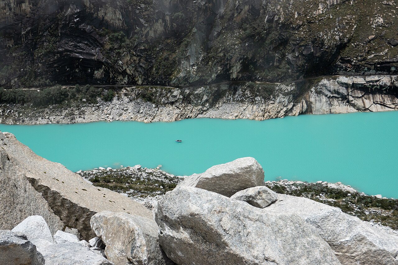 Turquoise glacial lake with a small boat below a pale rocky shoreline and dark cliffs in the Andes