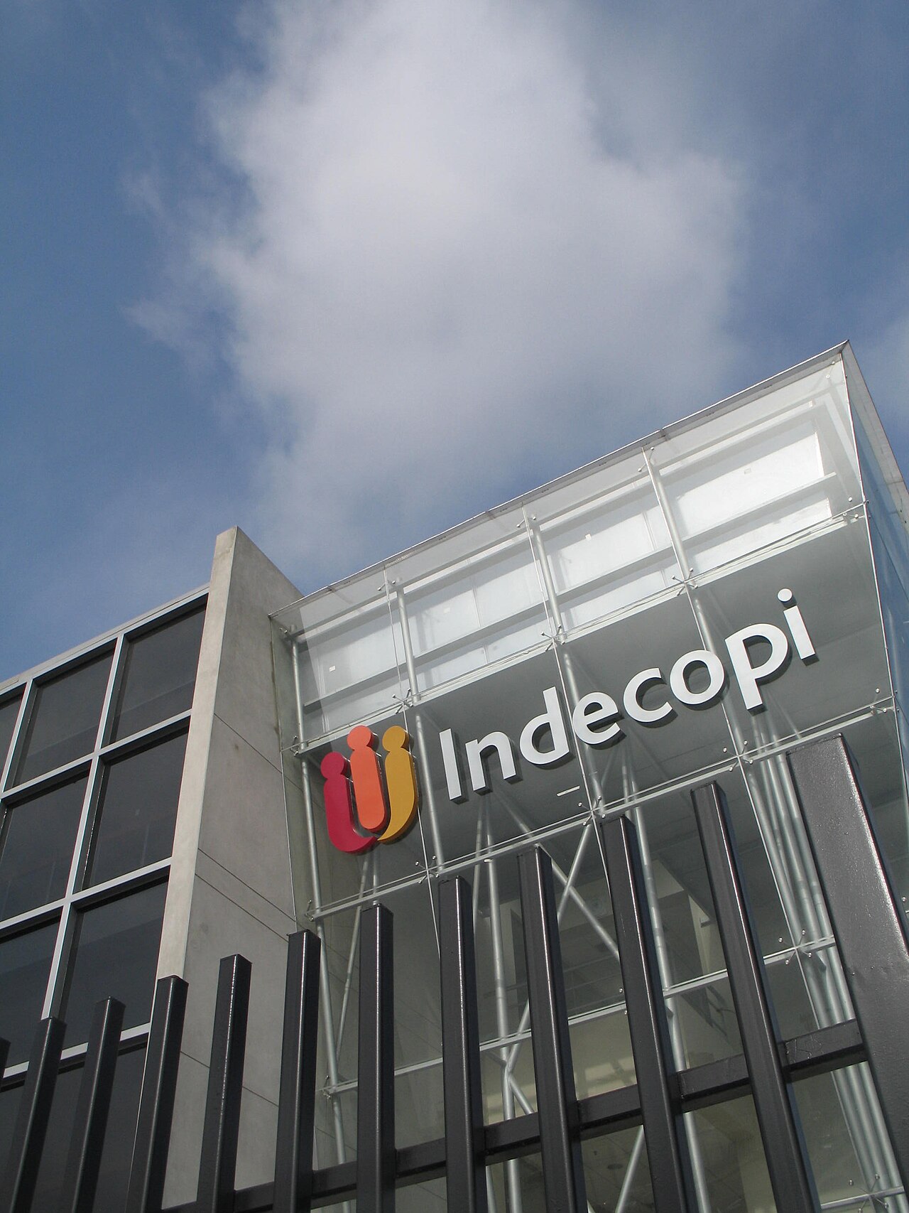 Glass facade of an Indecopi building with the agency's red, orange and yellow logo and name, under a blue sky