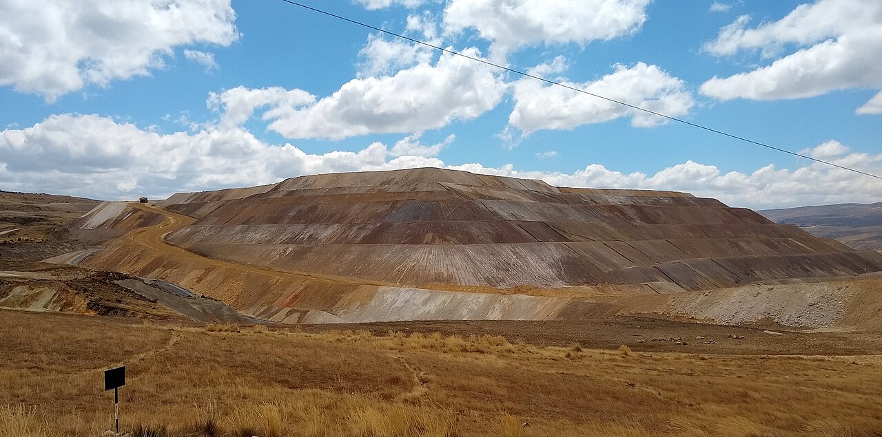 Brown terraced slopes at a large open-pit gold mine in the Peruvian highlands under a blue sky, with dry grassland in front