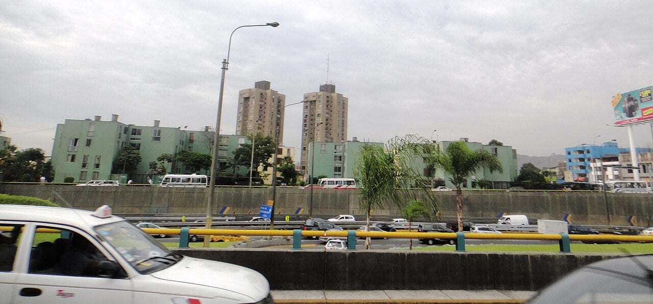 Cars and minibuses on a sunken highway in Lima with apartment blocks and two tall towers behind under an overcast sky