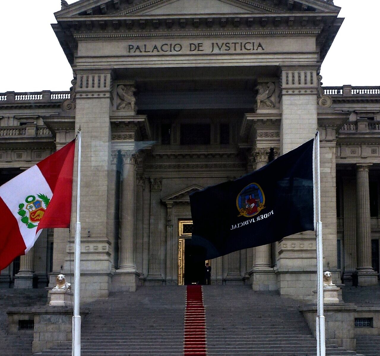 The stone facade of the Palace of Justice in Lima with the words Palacio de Justicia, a red carpet on the steps and the flags of Peru and the judiciary