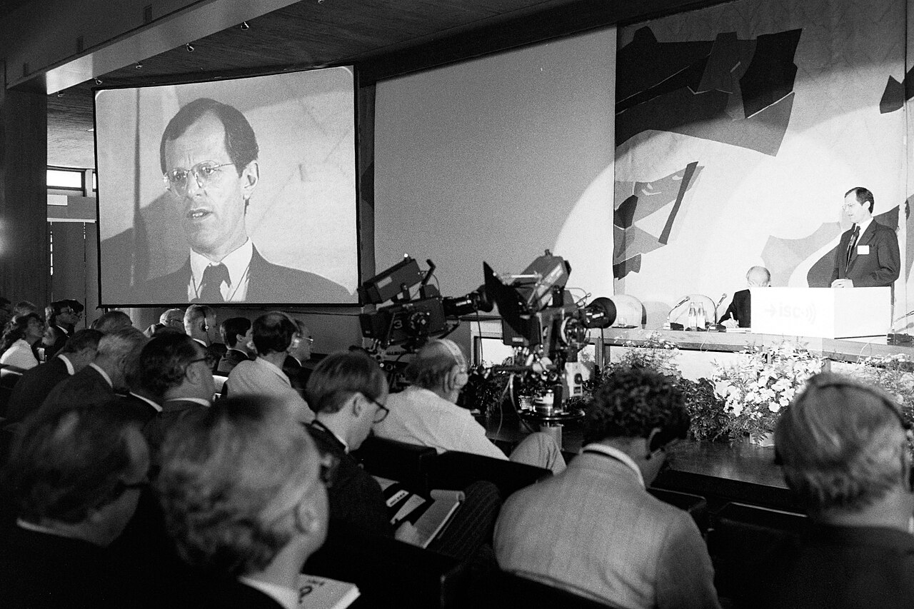 Black-and-white photo of a conference hall where Pedro Pablo Kuczynski appears on a large screen above an audience and television cameras