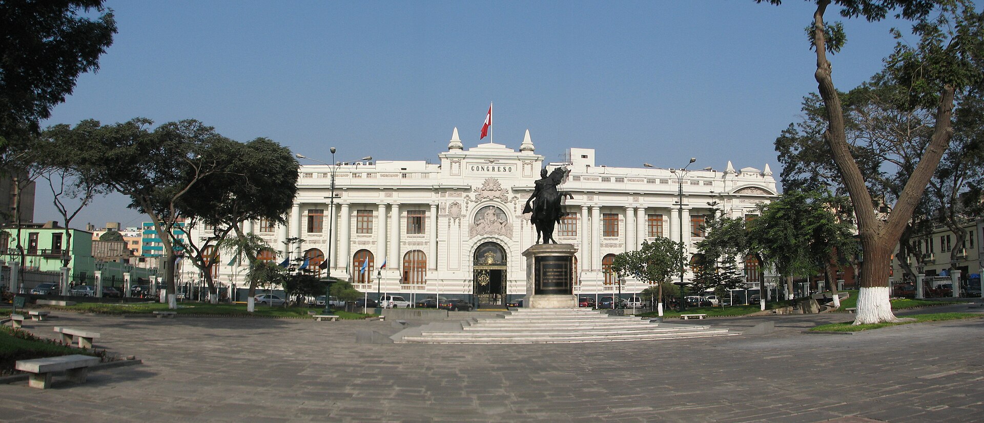 Wide view of Peru’s Congress building in central Lima behind the Simón Bolívar monument, with a Peruvian flag above the entrance, trees and a paved plaza