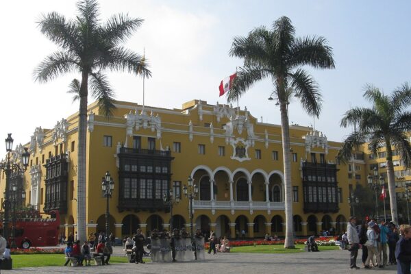 Lima's yellow Municipal Palace with wooden balconies on the Plaza de Armas, palm trees, a Peruvian flag and police officers in front