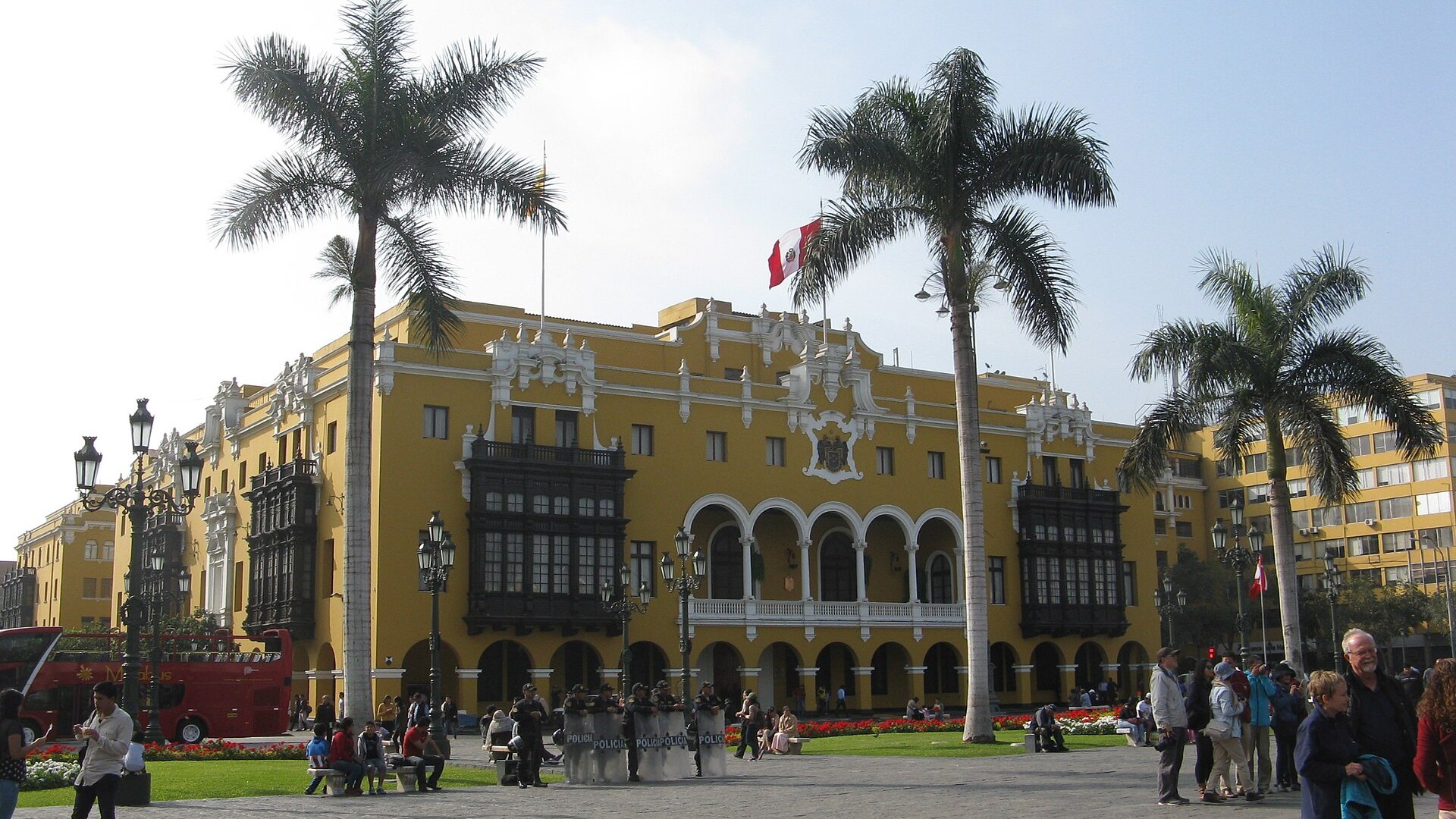 Lima's yellow Municipal Palace with wooden balconies on the Plaza de Armas, palm trees, a Peruvian flag and police officers in front