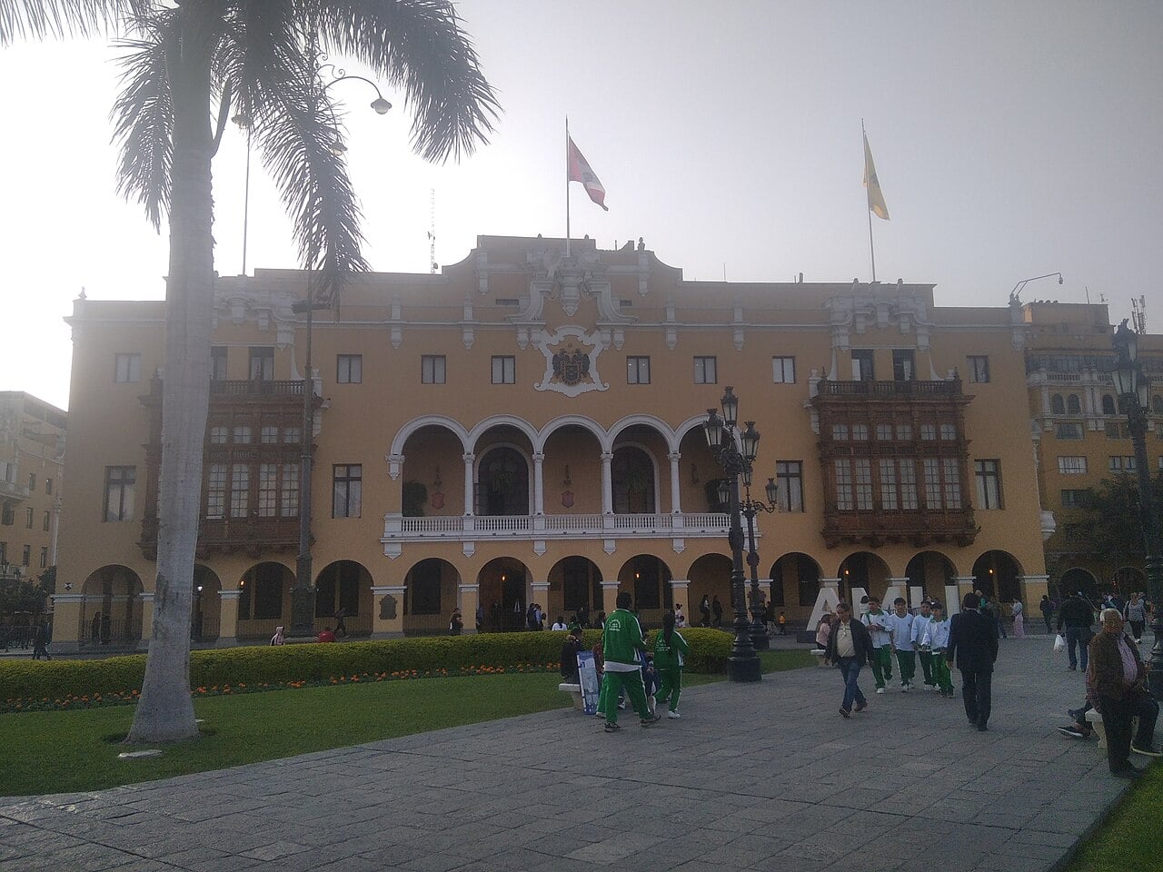 Yellow colonial-style facade of Lima's Municipal Palace with arched balcony and flags, seen from the Plaza Mayor