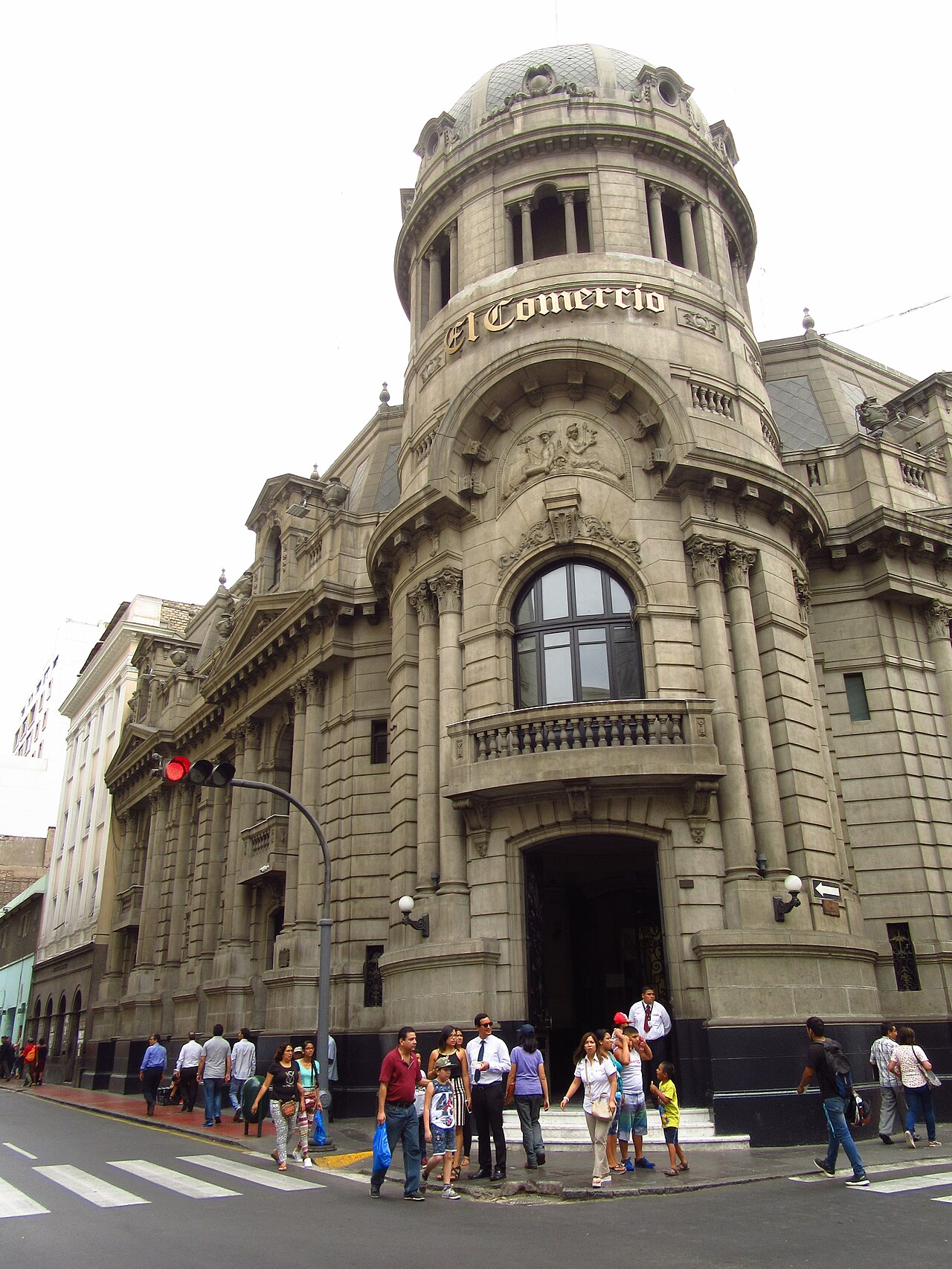 Pedestrians cross the street in front of the stone corner tower of the El Comercio building in Lima, its name in gold letters.