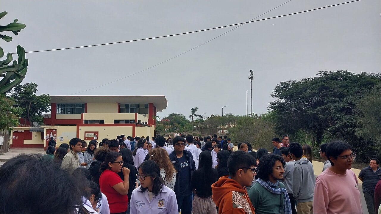 Crowd of university students in lab coats and jackets standing outside a red and cream campus building in Lima during an earthquake drill